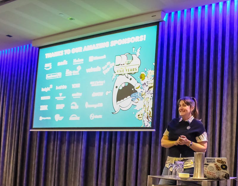 Kristy Sachse standing in front of her presentation at DDD Perth 2019. She is smiling at the camera. The presentation has a set of sponsor logos and an image of a shark with the text bubble “DDD Perth celebrating 5 years”