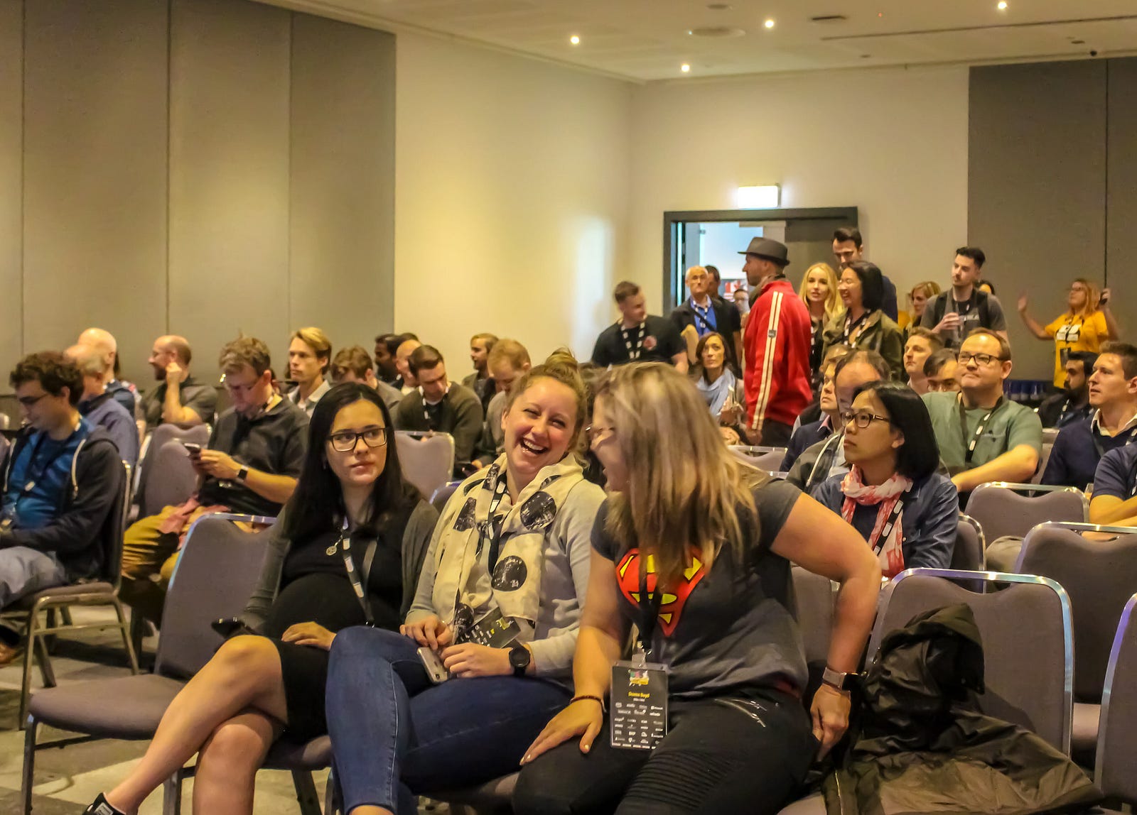People sitting in the front row of a room full of chairs. They are smiling and laughing. People behind them are standing and sitting, some are reading their phones.