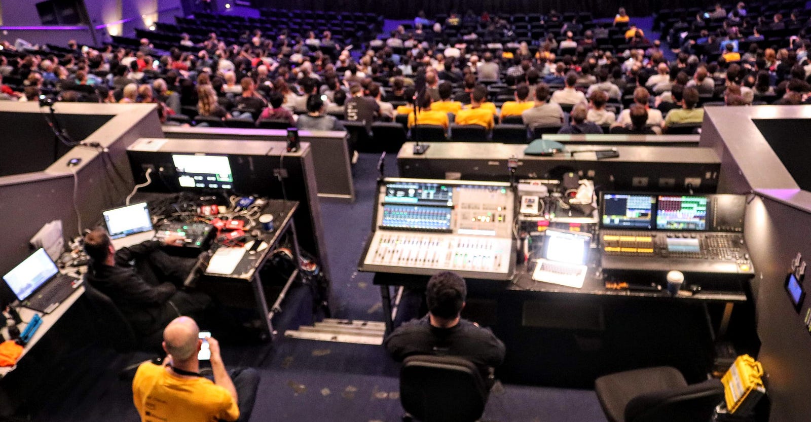 An almost birdseye view of the AV mixing desk in a large theatre. There are two people sitting in the space, and there are hundreds of people in front of them, seated in the theatre.