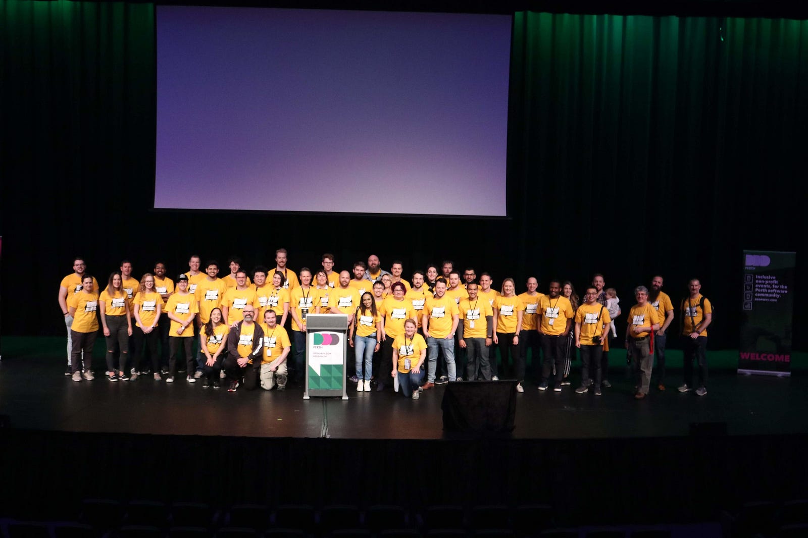 Over 50 people on a large stage all wearing yellow shirts that say “DDD Perth 2019 Team”. Some people in the front are kneeling. The majority of people are standing in multiple rows. There is a lectern in the front with a DDD Perth banner on it.