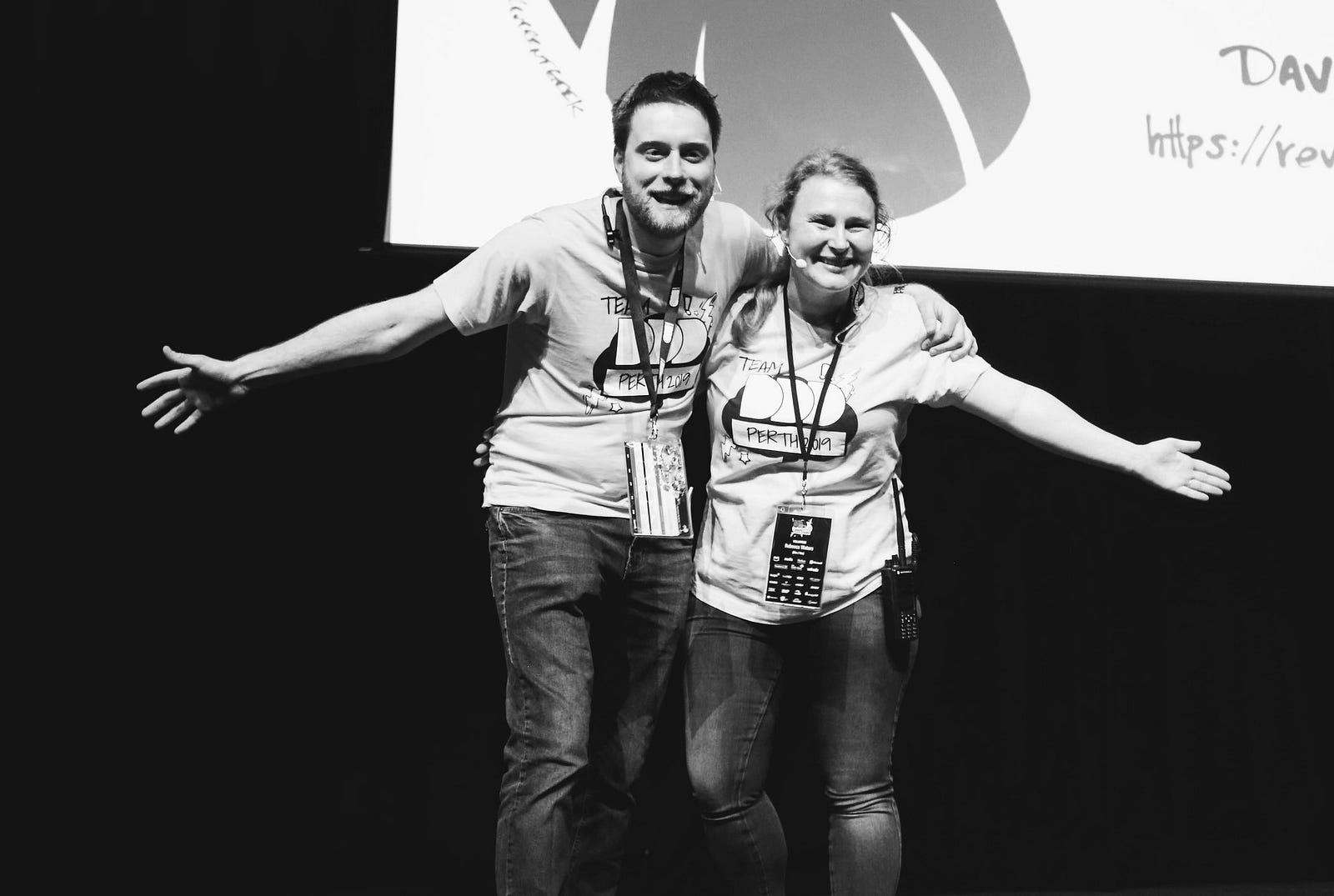 Two people on stage, hugging side on and facing the audience who are not visible. They have their arms wide open and are smiling. The photo is in black and white.